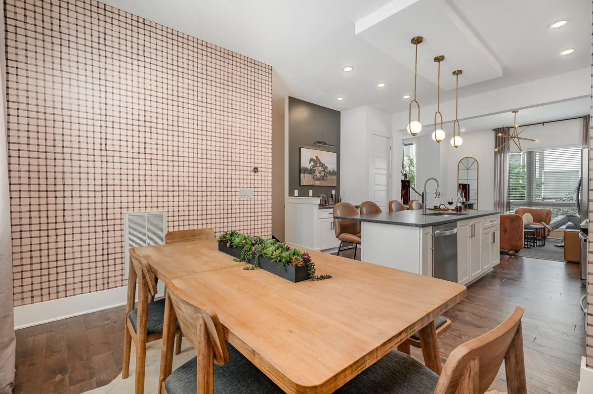This image presents a beautifully set dining table with a modern grid-patterned wall behind it, a kitchen island to the side, and stylish hanging light fixtures above. The open layout hints at the kitchen and living areas being closely connected, ideal for socialization.