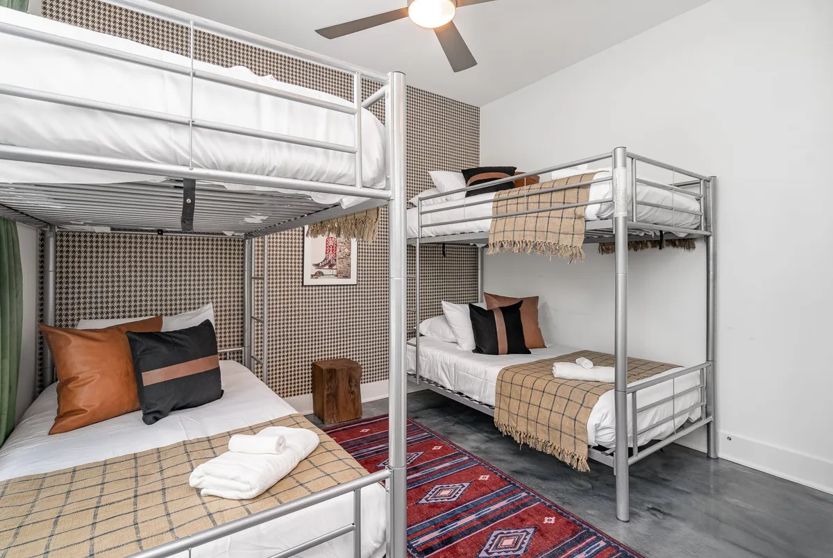 A playful bunk room with two sets of bunk beds, a colorful rug, textured wall, wooden stool, and decorative pillows. The space exudes a relaxed, kid-friendly vibe with plenty of room for play and late-night conversations.