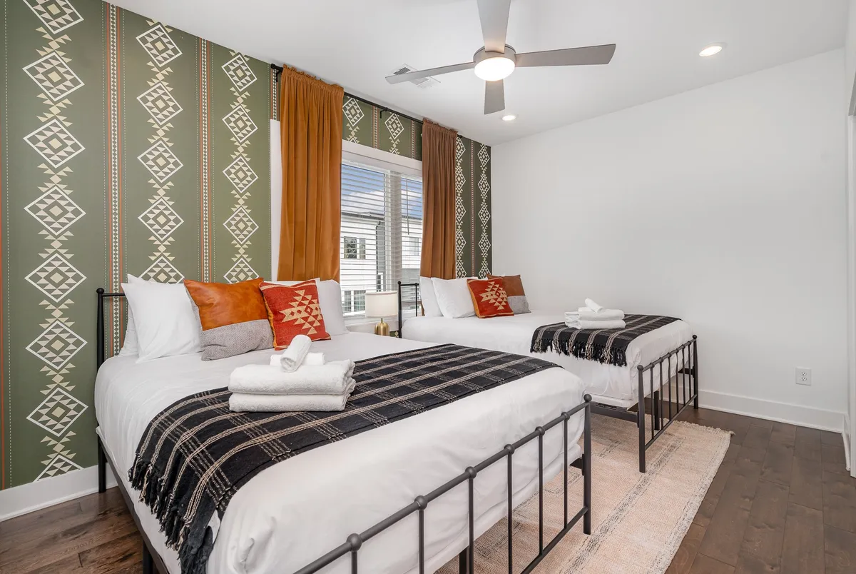 A spacious bedroom with two beds, each featuring decorative throws, backed by colorful patterned wallpaper and bathed in natural light from large windows. The space feels airy, comfortable, and ideal for friends or families.