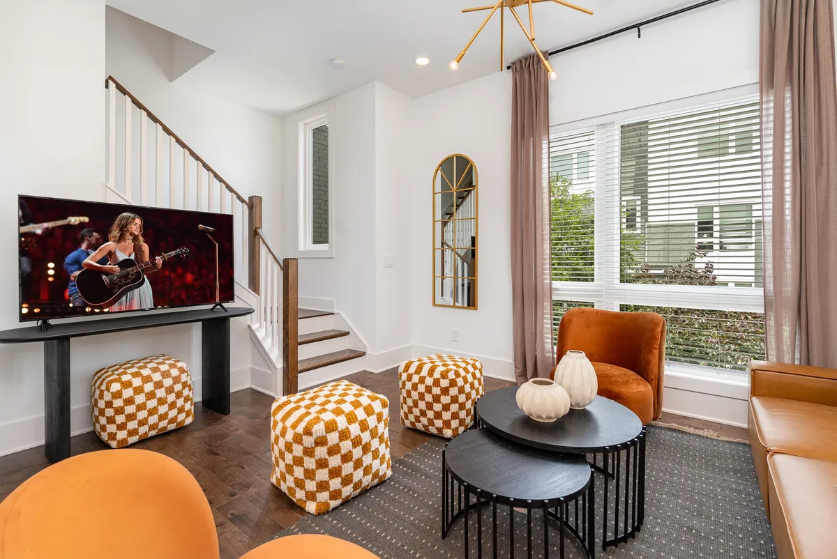 A living area with a plush brown leather sofa, round coffee table, and colorful poufs is oriented toward a large television. The space is filled with natural light from big windows, and the décor is contemporary yet comfortable.